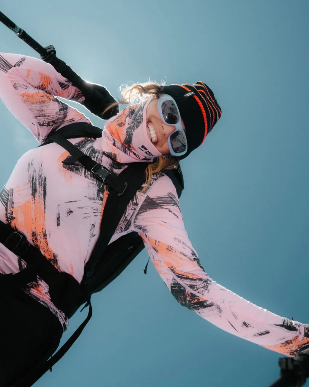 A smiling skier in sunglasses and a black beanie stretches out an arm against a blue sky.