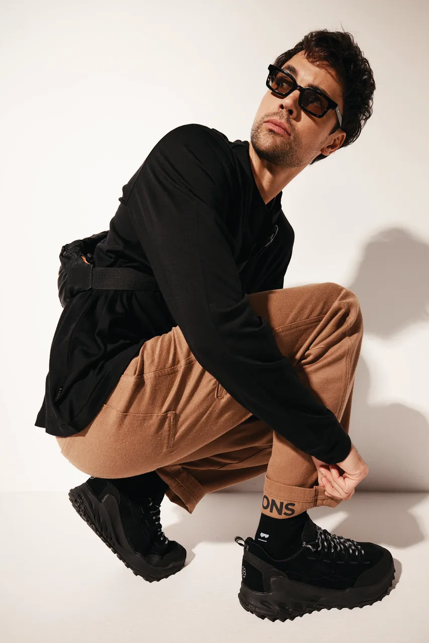 Model in a black jacket and tan trousers crouches in black trail shoes and socks.