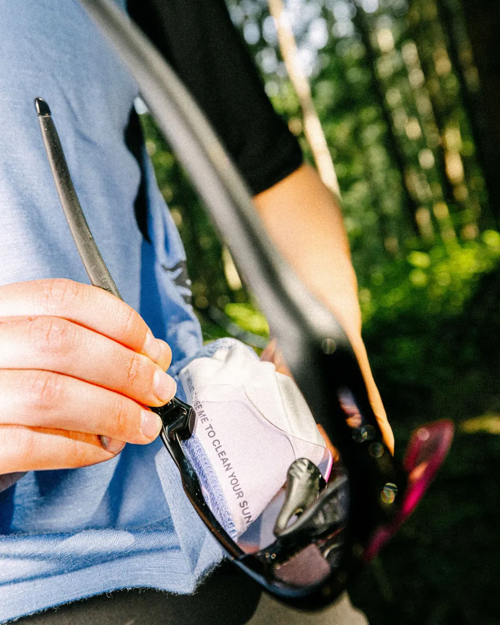 A person wipes sunglasses with a cloth outdoors in a sunlit forest.