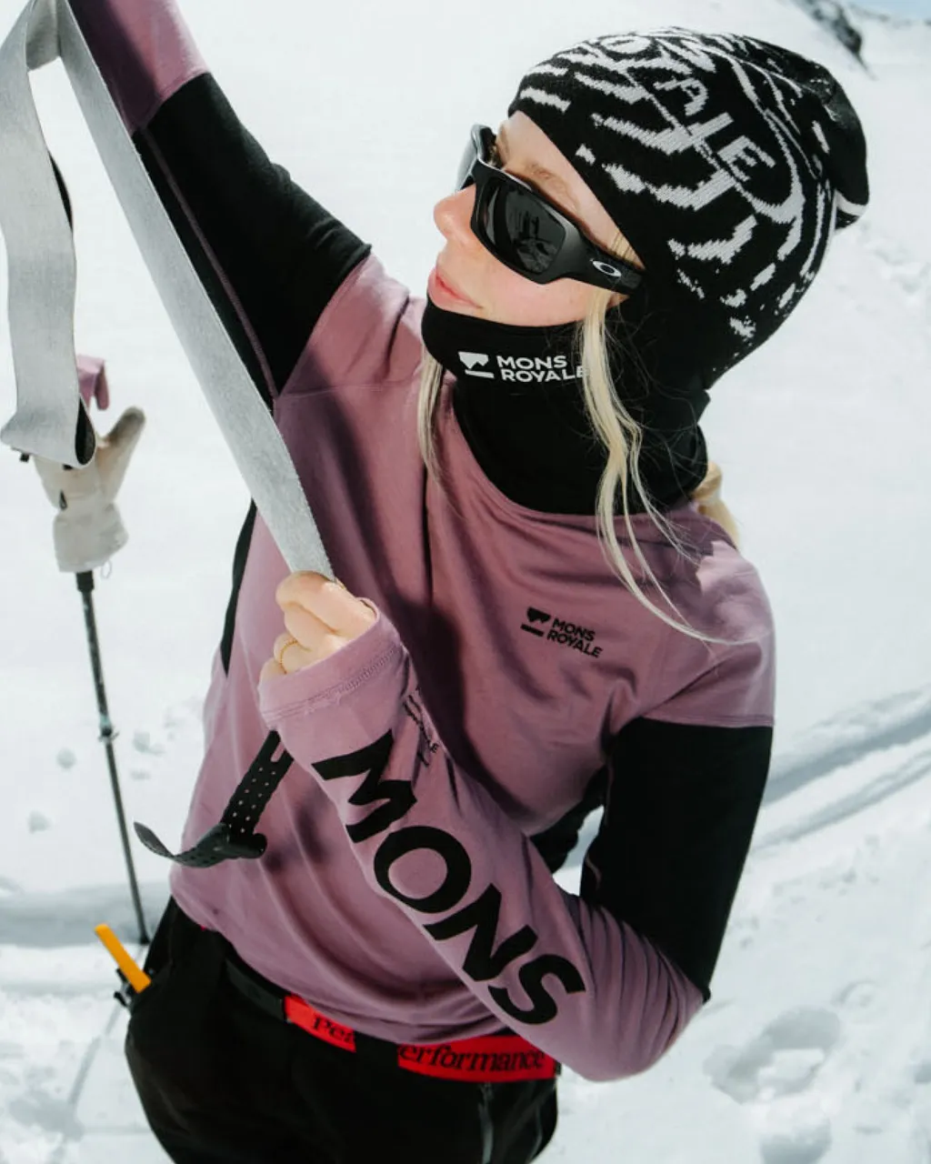 A skier in a mauve Mons Royale top and patterned beanie adjusts gear on a snowy slope.