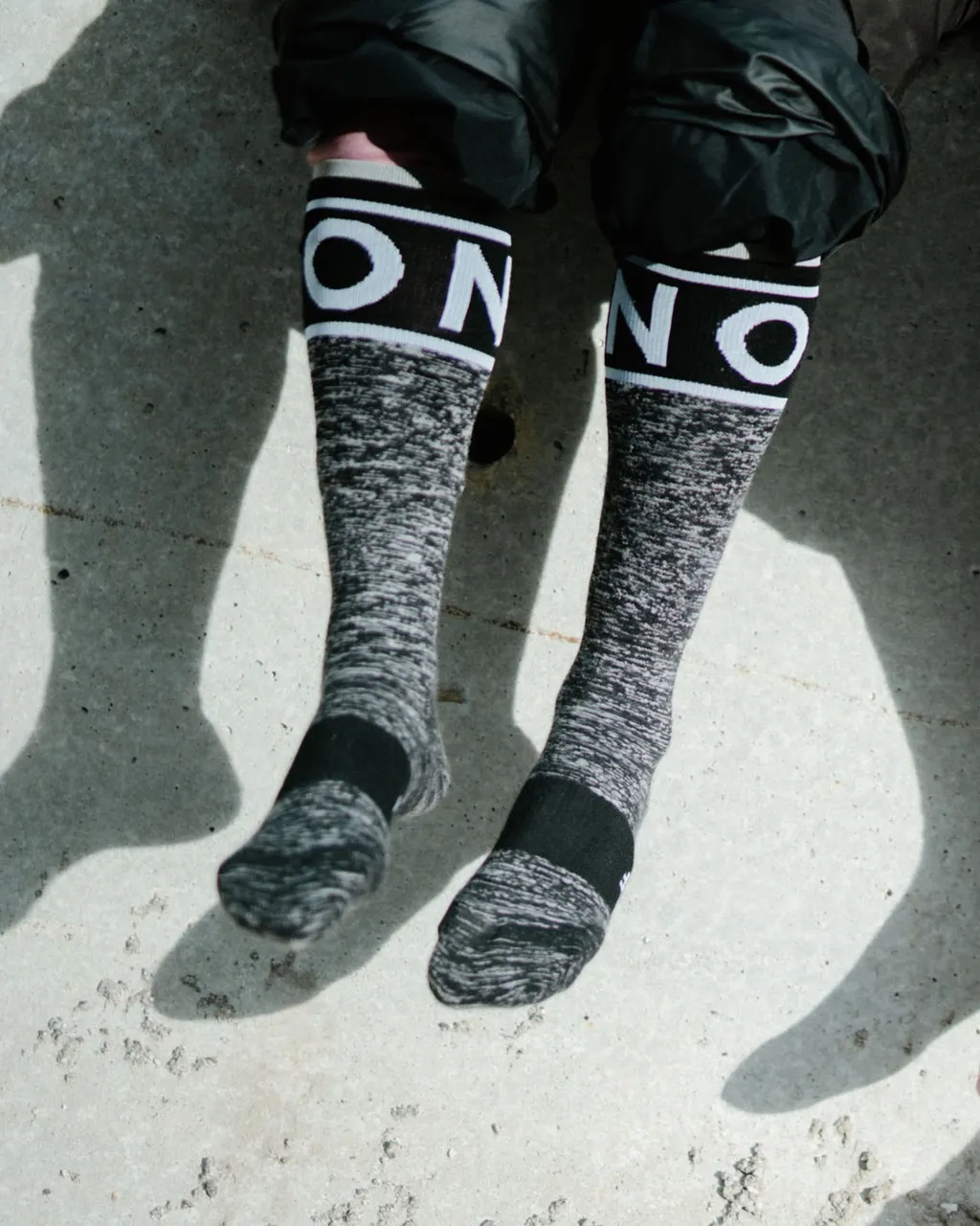 A person wears black and grey knee-high socks with bold logo cuffs on sunlit concrete.