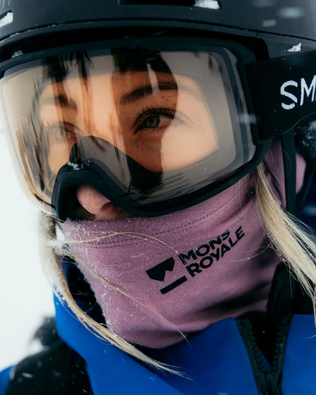 A skier wears black goggles and a purple Mons Royale neck warmer over a blue jacket.