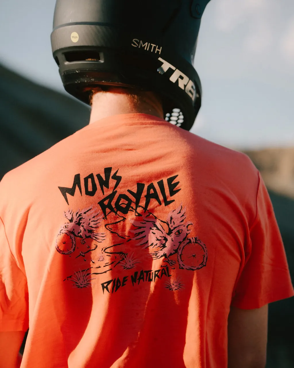 A person in a black helmet wears a coral Mons Royale T-shirt with a mountain-inspired back graphic.