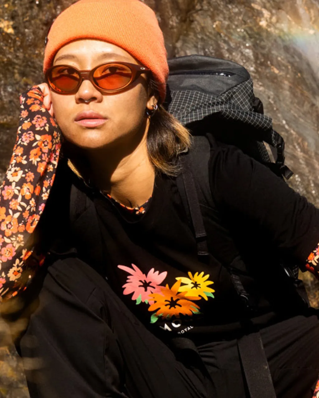 A person in an orange beanie and amber sunglasses wears a floral graphic shirt and backpack outdoors.