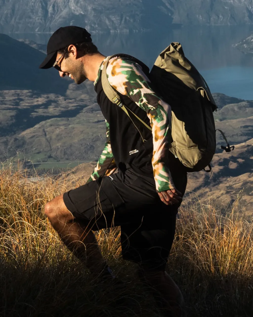 A hiker with a khaki backpack climbs through golden alpine grass against a mountain valley.