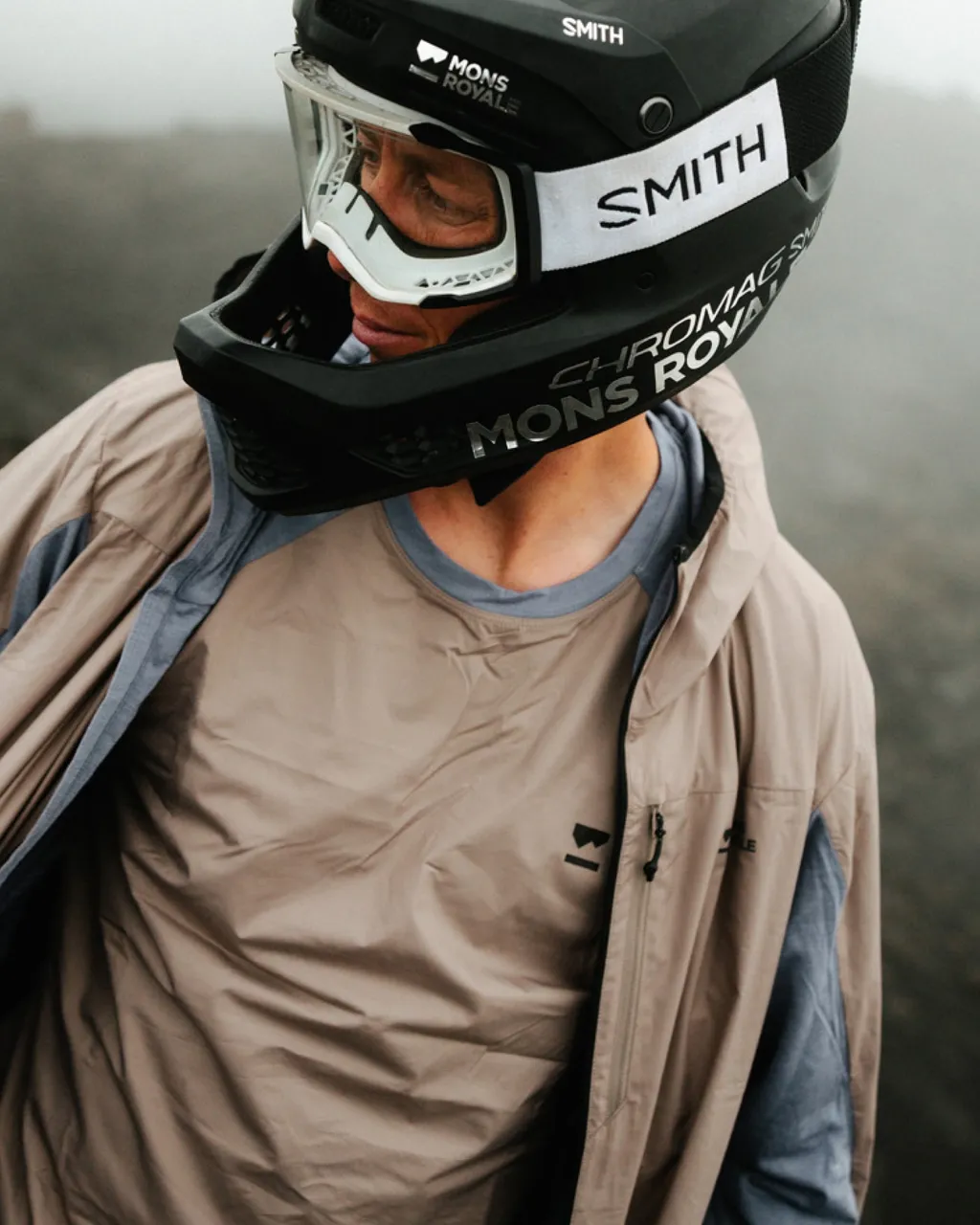 A rider in a branded black helmet and goggles wears a beige jacket over a grey shirt.