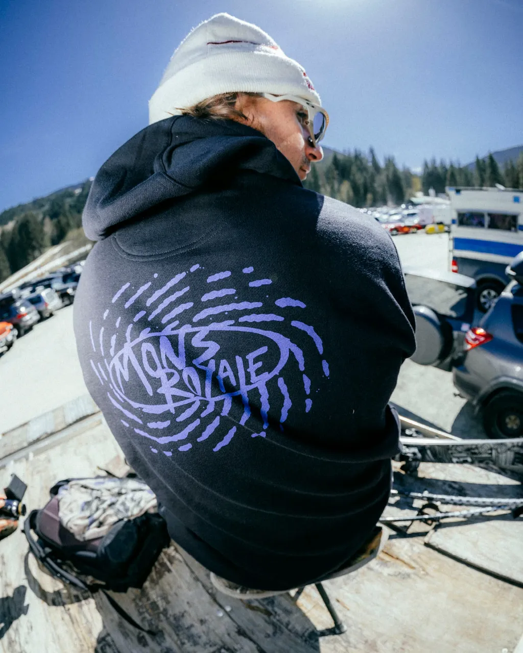 A person in a black Mons Royale hoodie and white beanie sits overlooking a mountain resort parking lot.