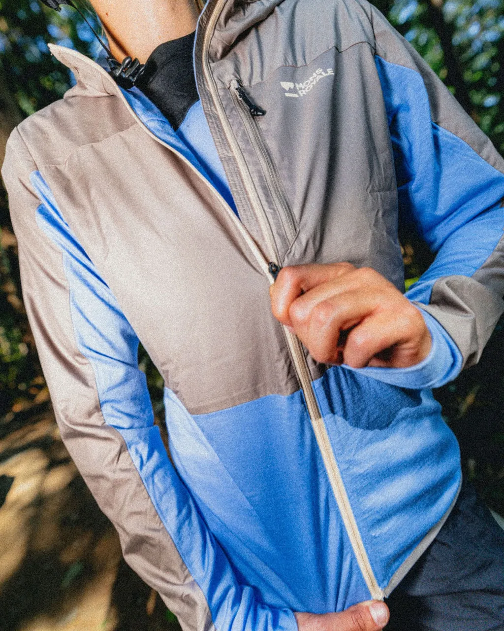 A person zips up a blue and grey colour-block Mons Royale jacket outdoors.