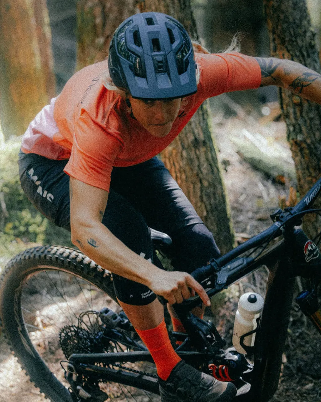 A mountain biker in an orange jersey rides a black bike along a wooded trail.