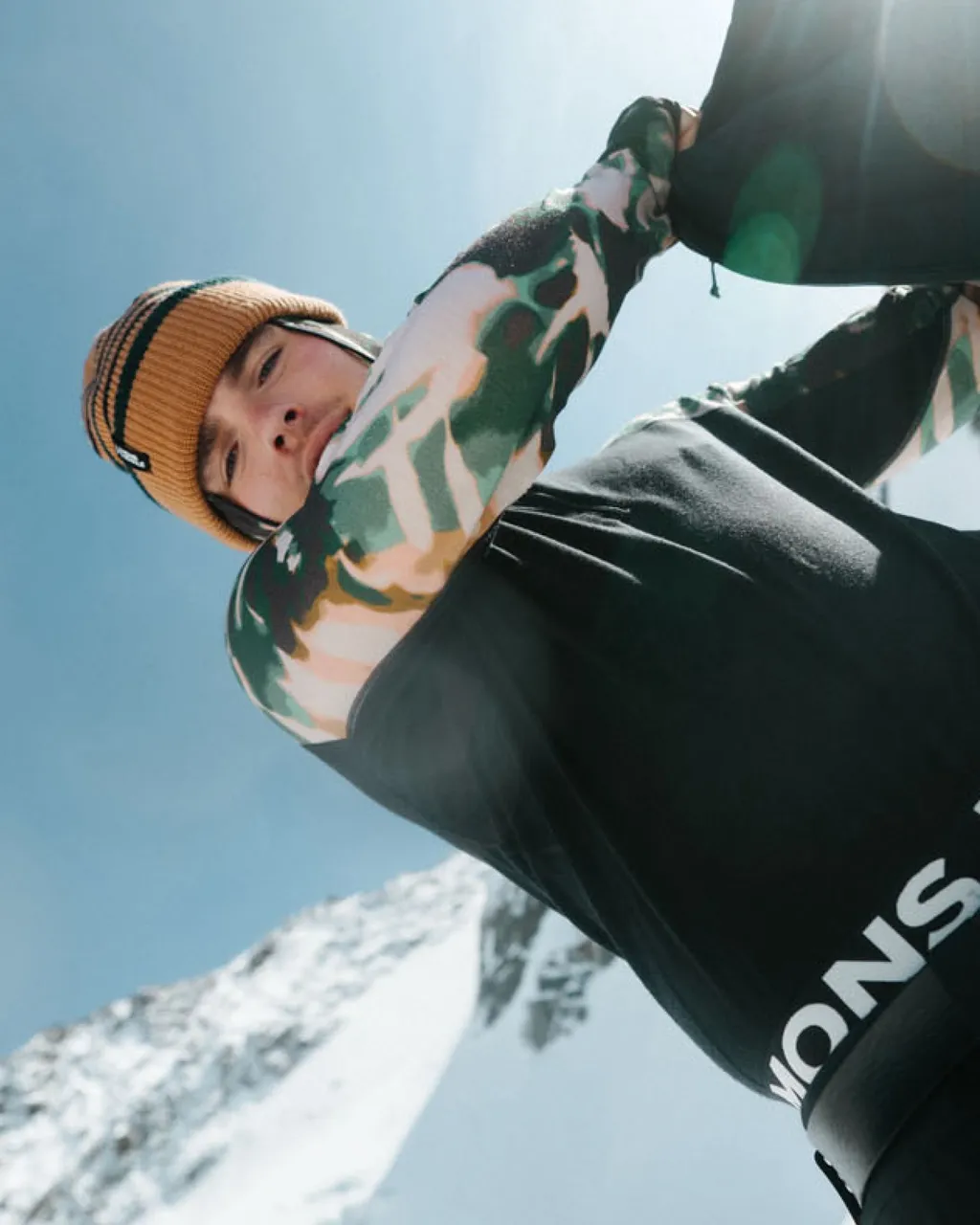 Snowboarder in a mustard beanie adjusts gear against a bright blue sky and snowy mountain.