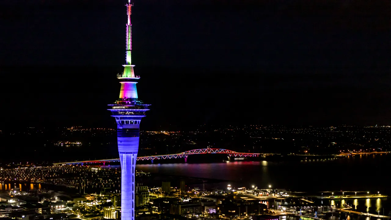 Sky Tower lit up rainbow coloured with Auckland Harbour Bridge light show in background