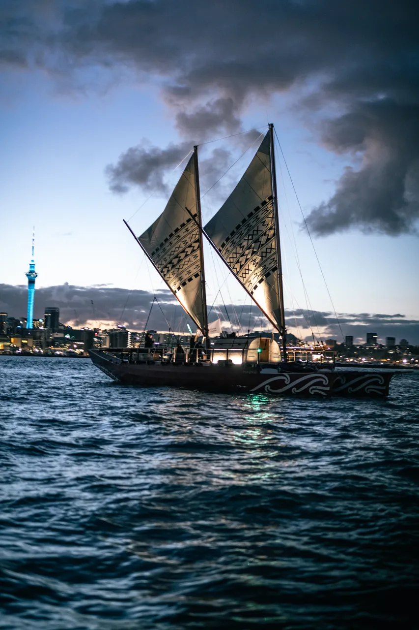 Waka Hourua sails The Waitematā Harbour as the Sky Tower is lit up blue