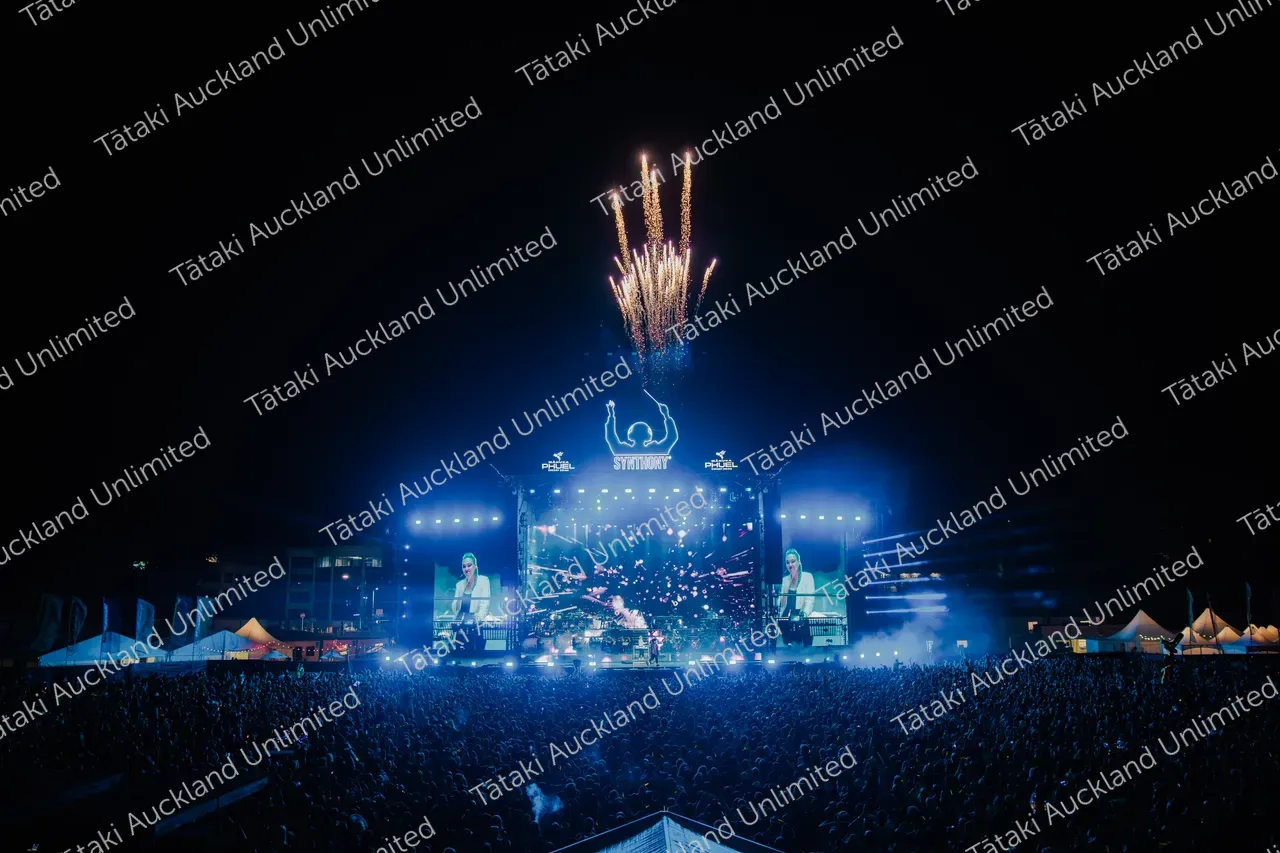 Fireworks burst above a brightly lit outdoor concert stage as a large crowd watches.