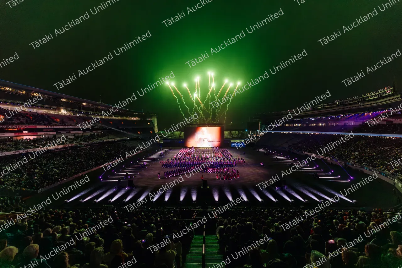 Green fireworks burst above performers on a stadium field as a large crowd watches.