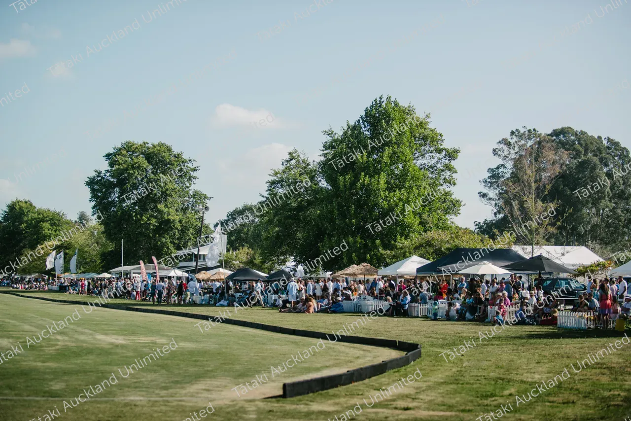 People gather by a grassy field at an outdoor event lined with tents and stalls.