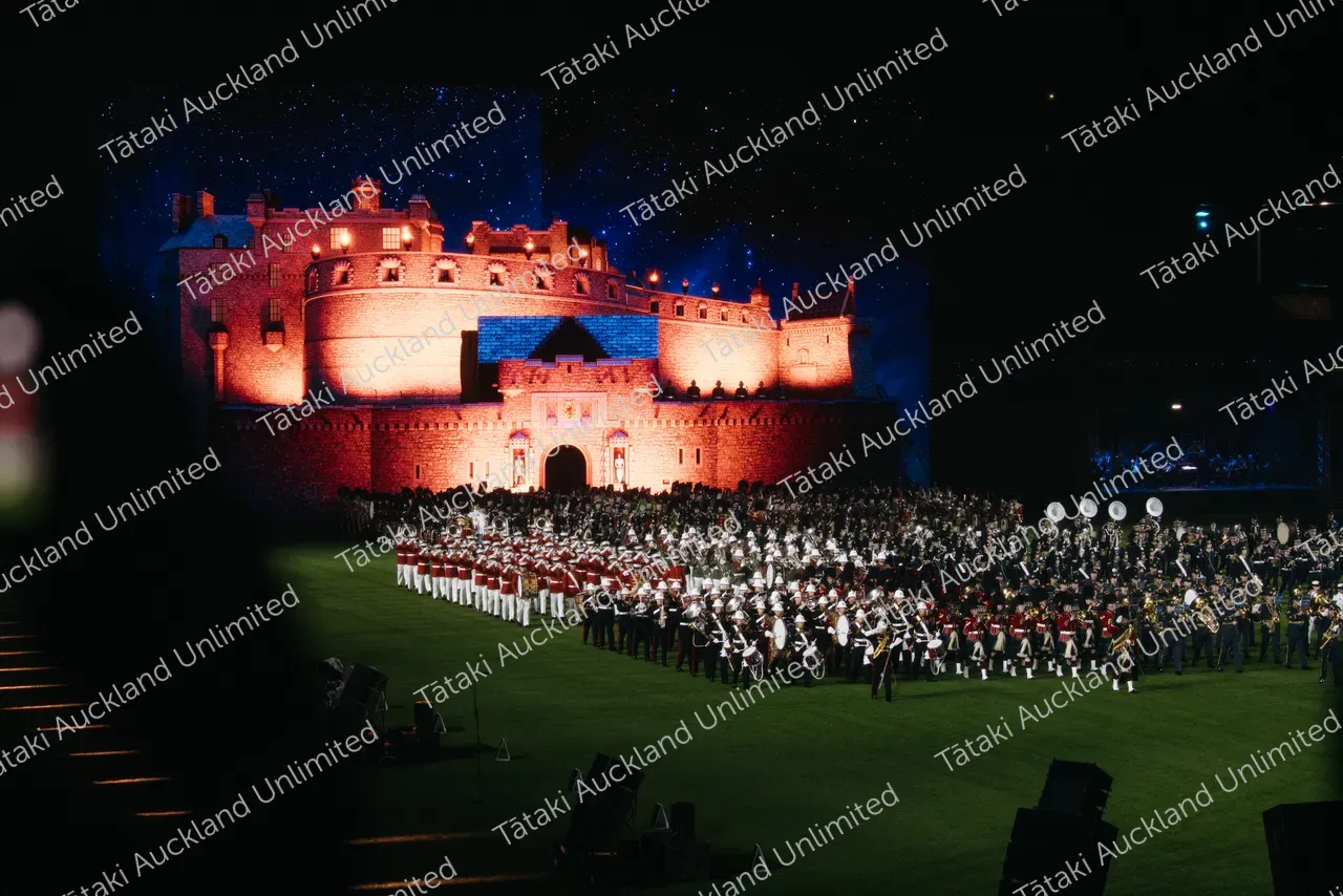 A marching band performs on a field before a brightly lit castle at night.