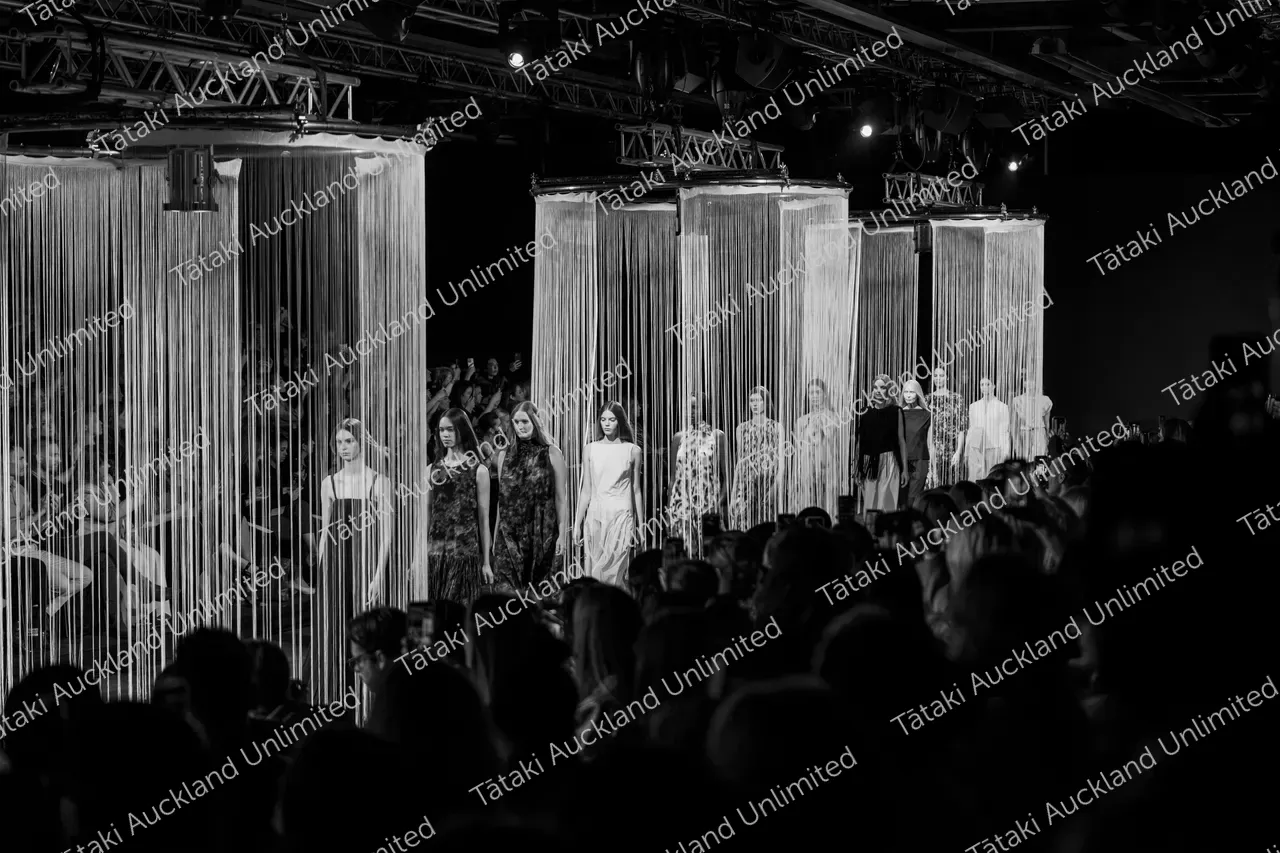 Models walk through hanging string curtains on a runway as the audience watches.