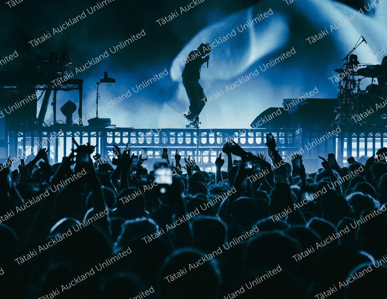 A silhouetted performer takes the stage as a crowd raises their hands under blue concert lights.