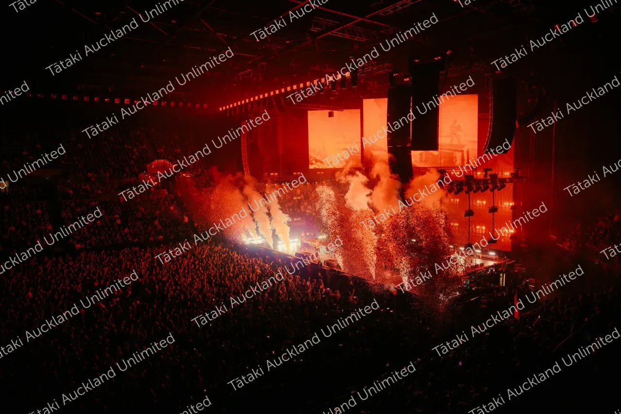 Red lights and smoke jets fill a concert arena as a large crowd watches the stage.
