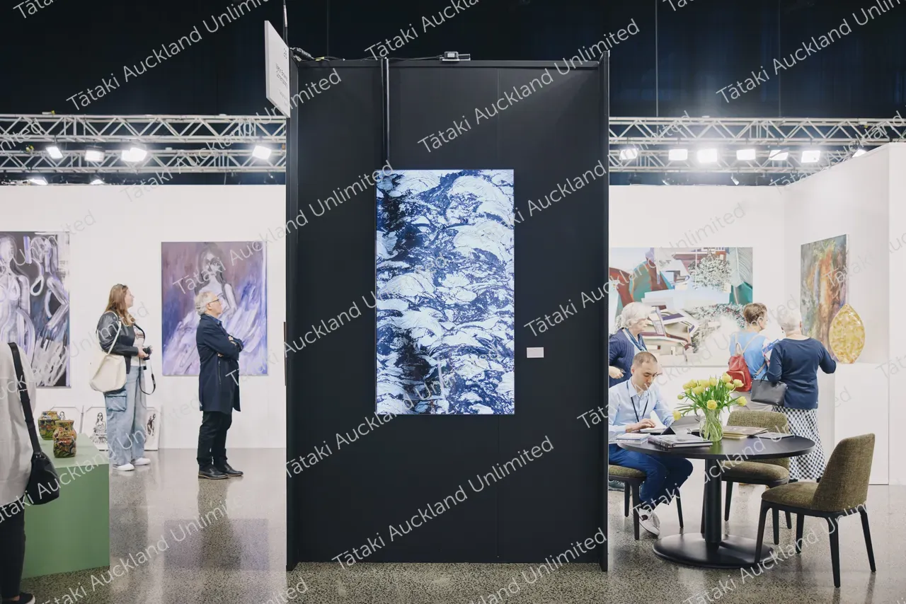 Visitors browse contemporary paintings at an art fair, including a blue-and-white abstract work in a black booth.