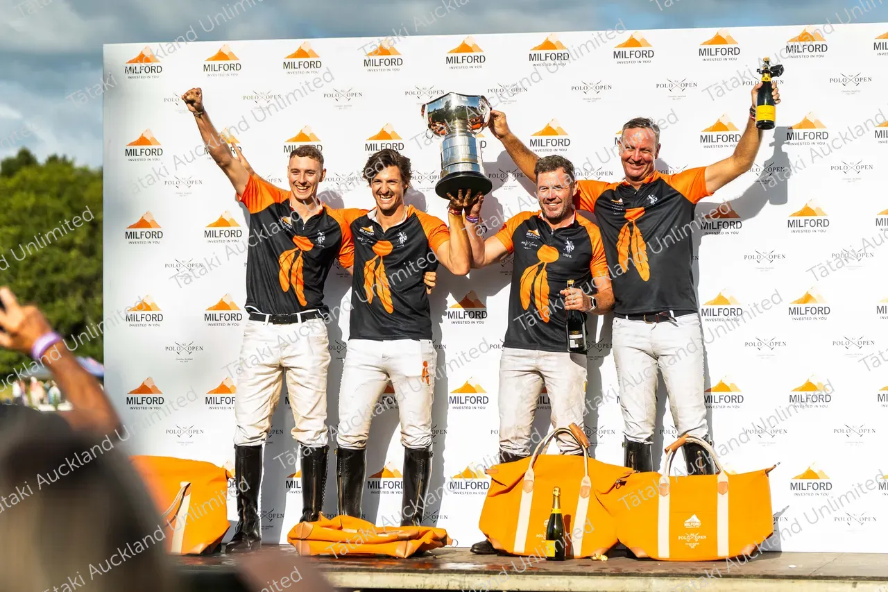 Four sailors in black and orange celebrate on a podium, raising a silver trophy and a bottle.