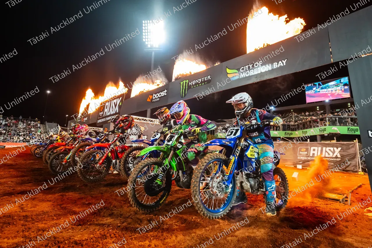 Motocross riders line up on dirt bikes beneath flames and floodlights at a night event.