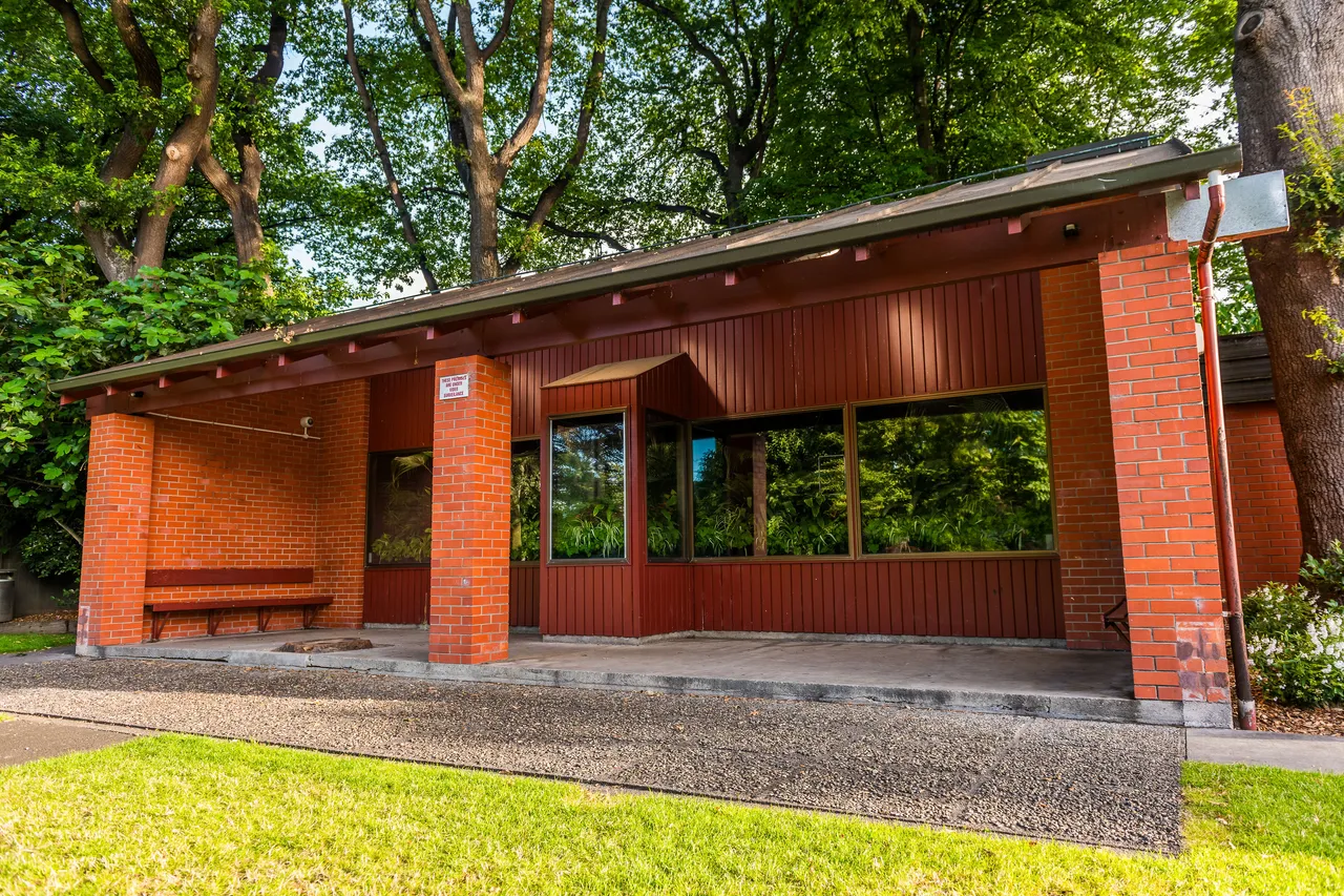 Small red brick park building with a bench under a canopy, surrounded by trees.