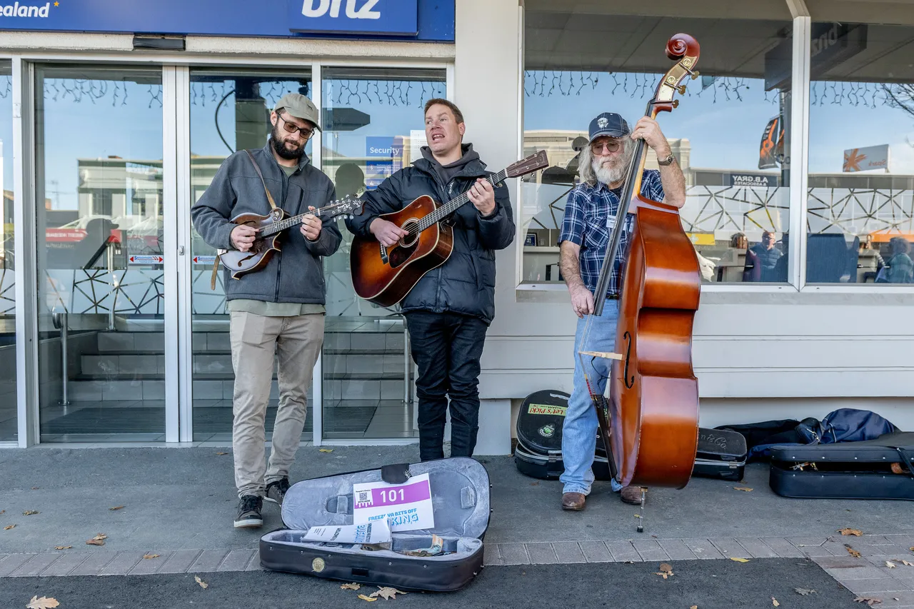 Three men busking. One playing a ukulele, one a guitar and one a cello.