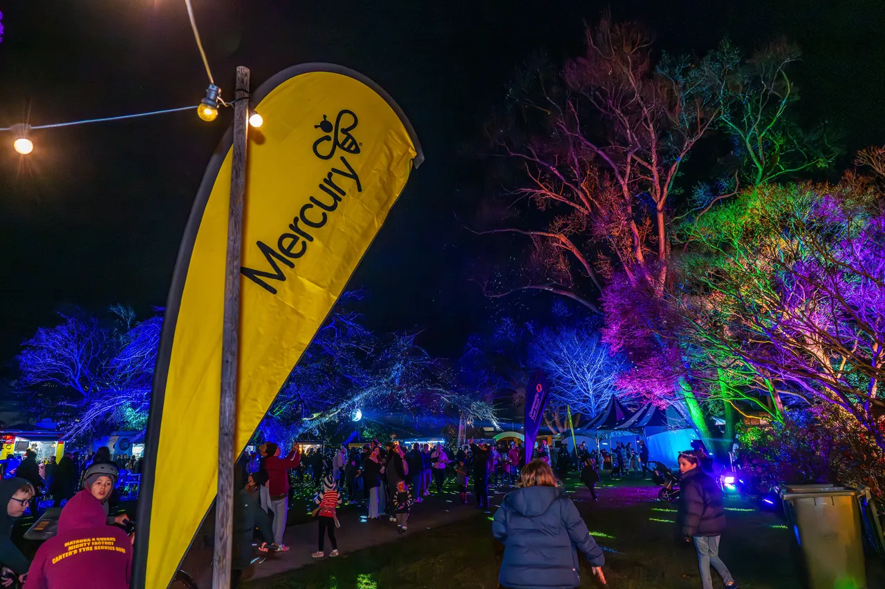 Mercury, sponsor's flag with crowd of people in background. Illuminated multi coloured trees.