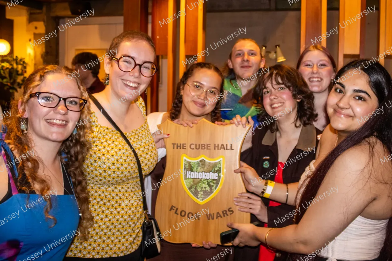 A group of students smile together while holding a wooden award plaque at an indoor event.