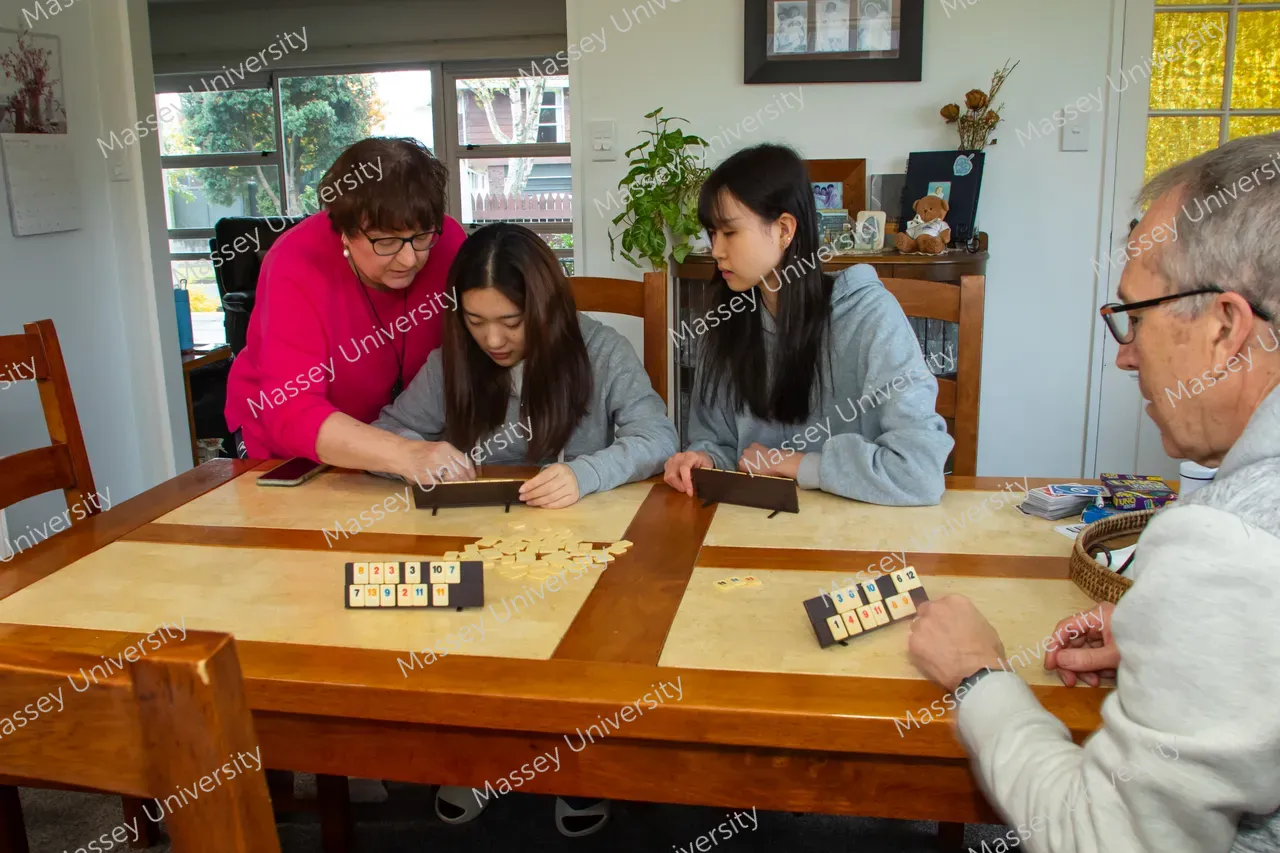 An older woman helps two young women with a tile-based board game as an older man sits across the table.