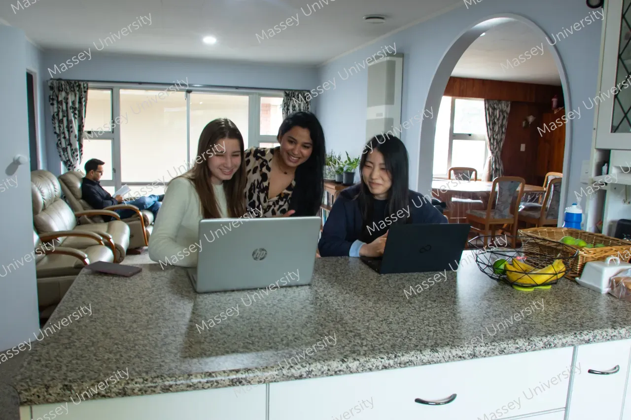 Three women work together on laptops at a kitchen counter while another person relaxes in the living room.