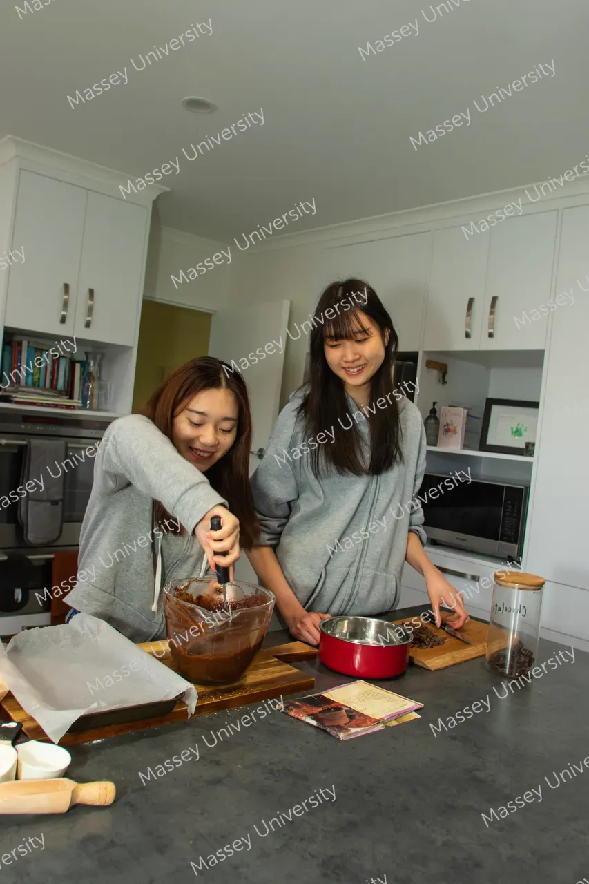 Two women prepare chocolate in a kitchen, stirring a bowl beside a red saucepan.