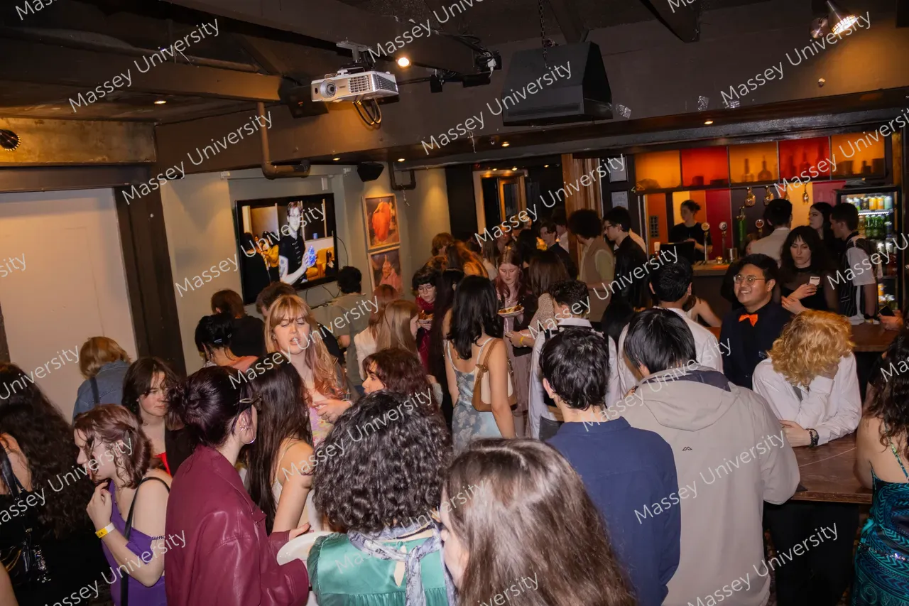 A crowd of people mingles and talks at a dimly lit indoor event beside a bar.