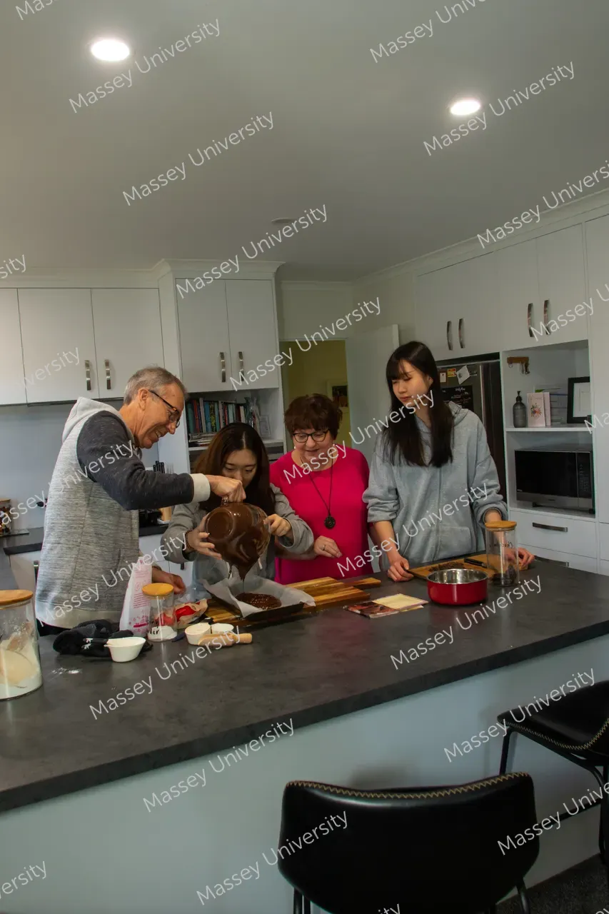 Four people prepare food together around a kitchen island, pouring ingredients from a bowl beside a pot.