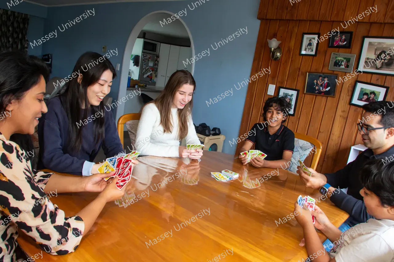 Six young people play a card game around a wooden table in a living room.