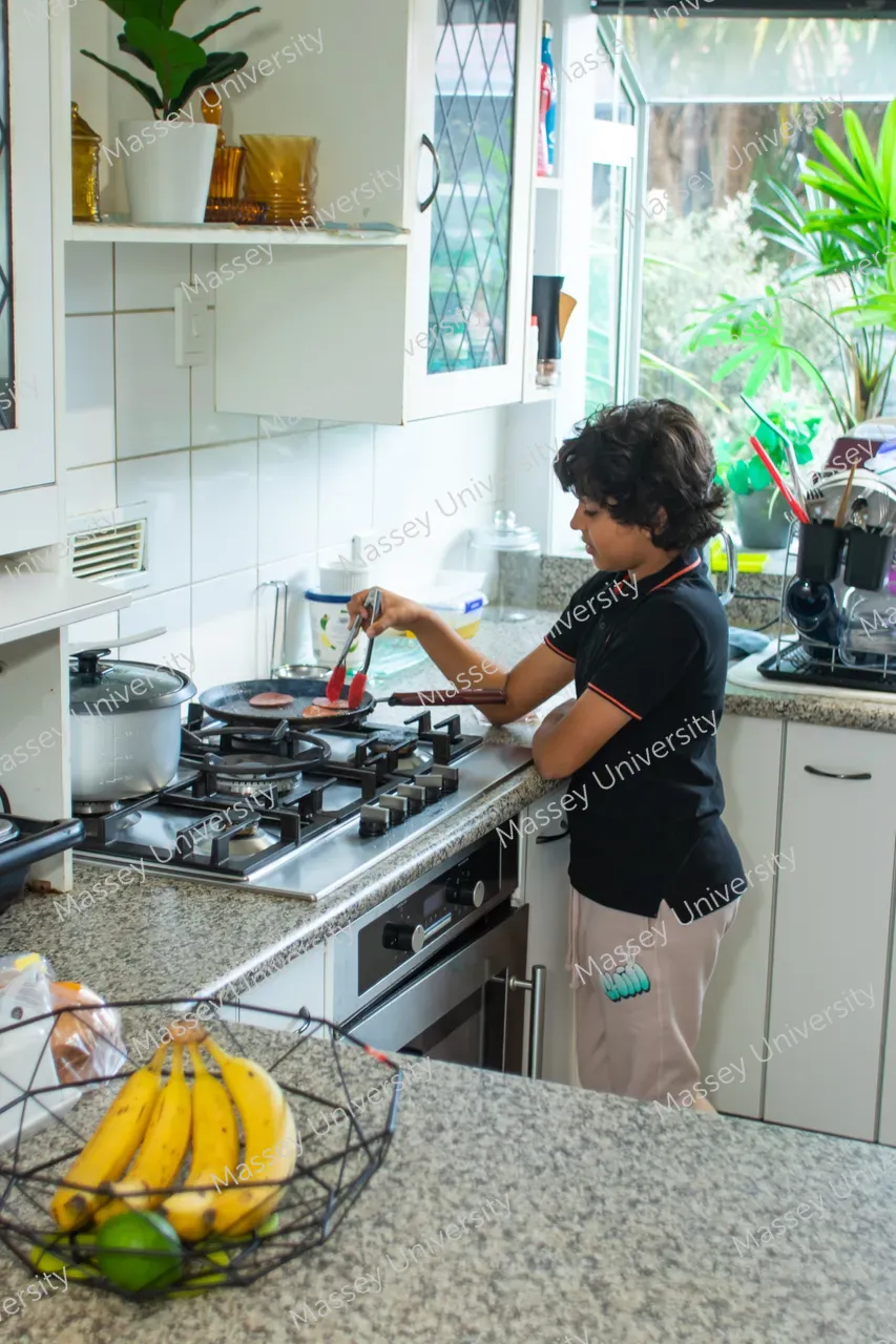 A person cooks at a gas stove while bananas and limes sit in a wire basket on the counter.