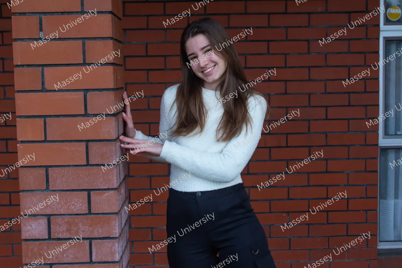 A smiling woman in a white sweater and black trousers leans against a brick wall.