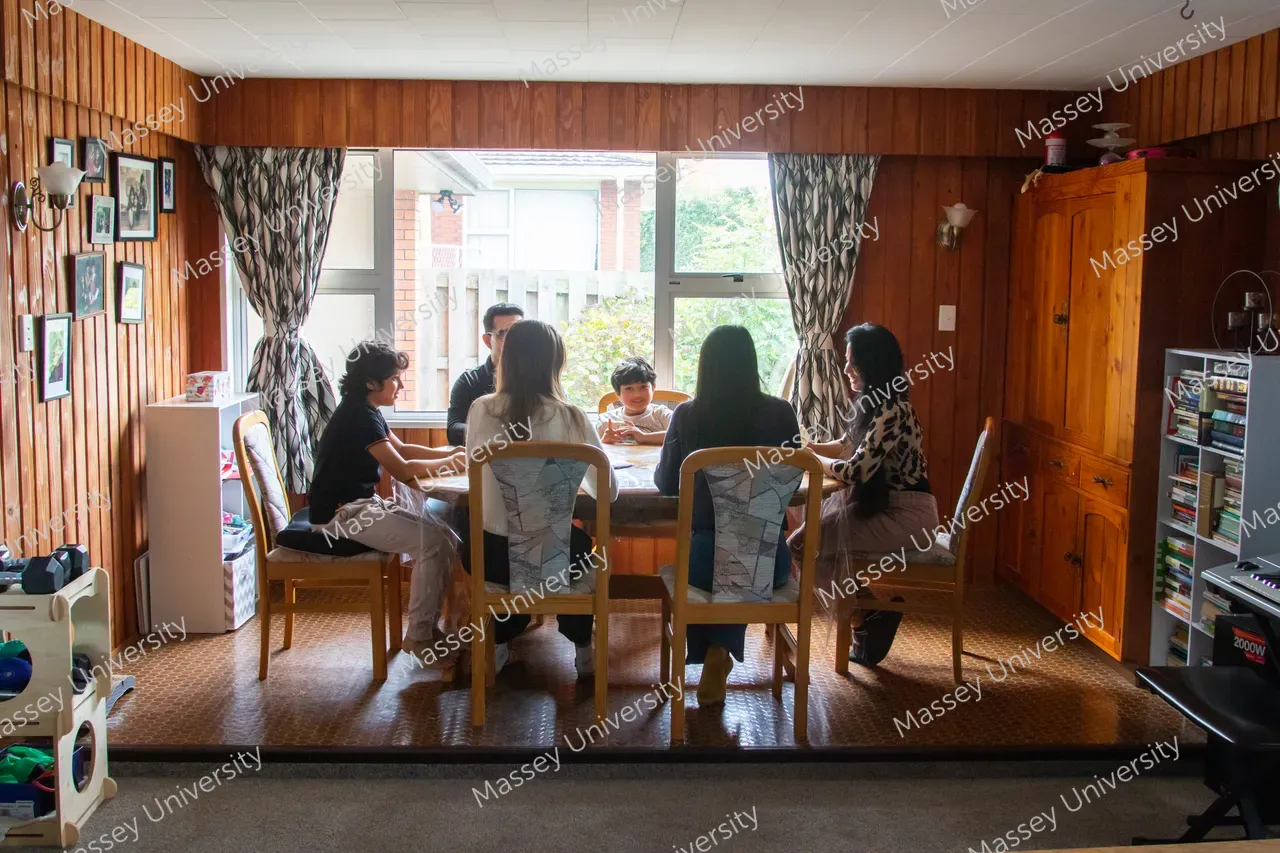 A family sits around a dining table in a wood-panelled room with a large window behind them.