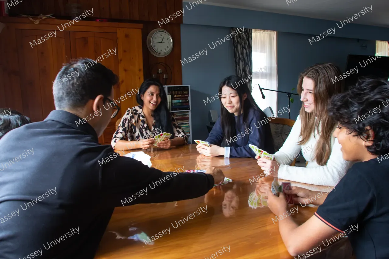 Five students sit around a wooden table, smiling as they play cards.