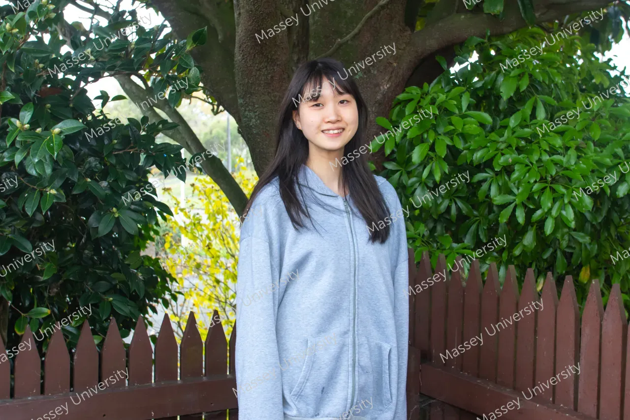 A smiling student in a grey hoodie stands beside a wooden picket fence under a leafy tree.