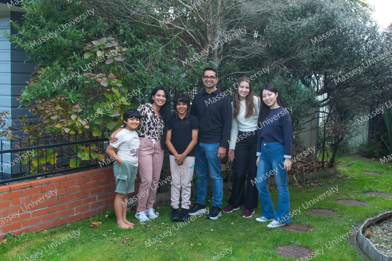 Six people pose together on a lawn beside a brick wall and leafy trees.