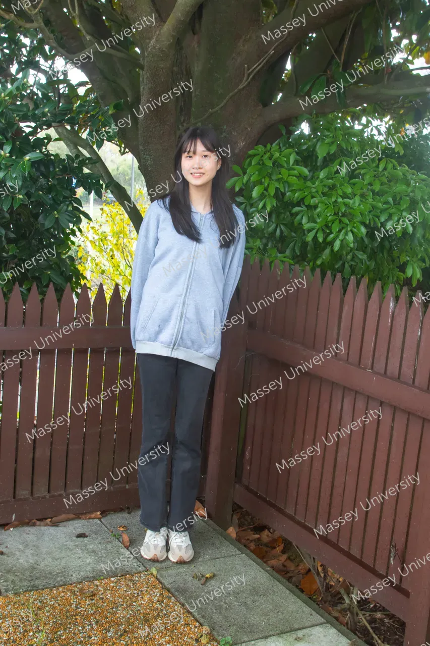 A young woman stands beneath a large tree beside a wooden fence in a garden.