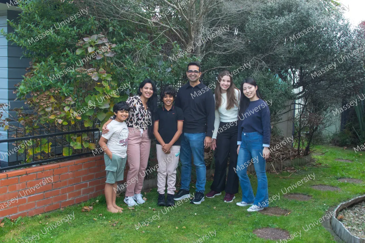 Six people stand together on a lawn beside a brick garden wall and trees.