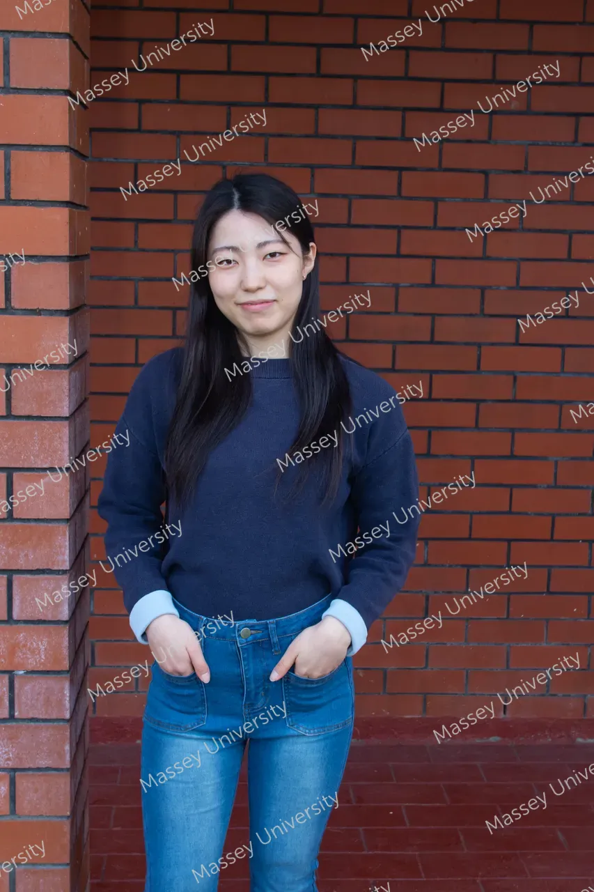 A young woman in a navy sweater and blue jeans stands against a red brick wall.