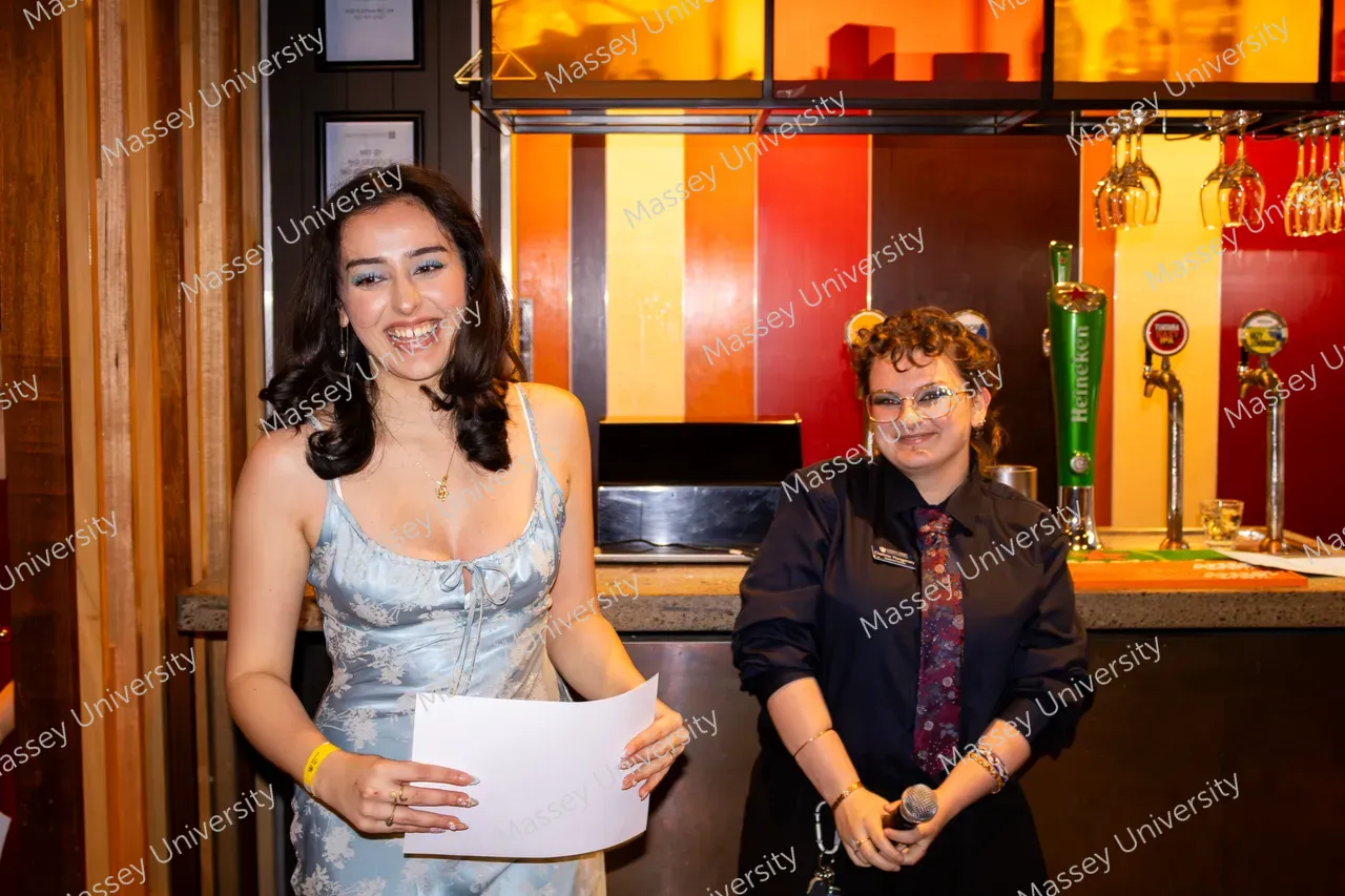 Two people smile at a venue, one holding a sheet of paper and the other a microphone beside a bar.