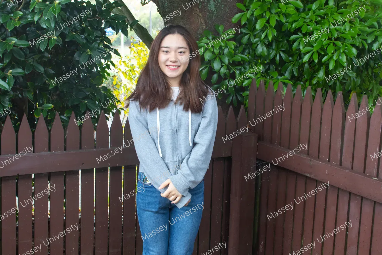 A smiling woman in a grey hoodie and jeans stands by a wooden fence among leafy shrubs.