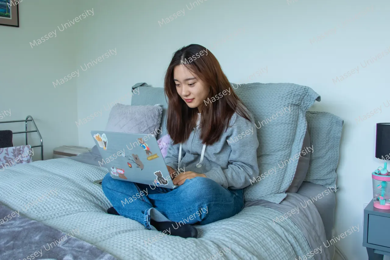 A student sits cross-legged on a bed and uses a sticker-covered laptop.