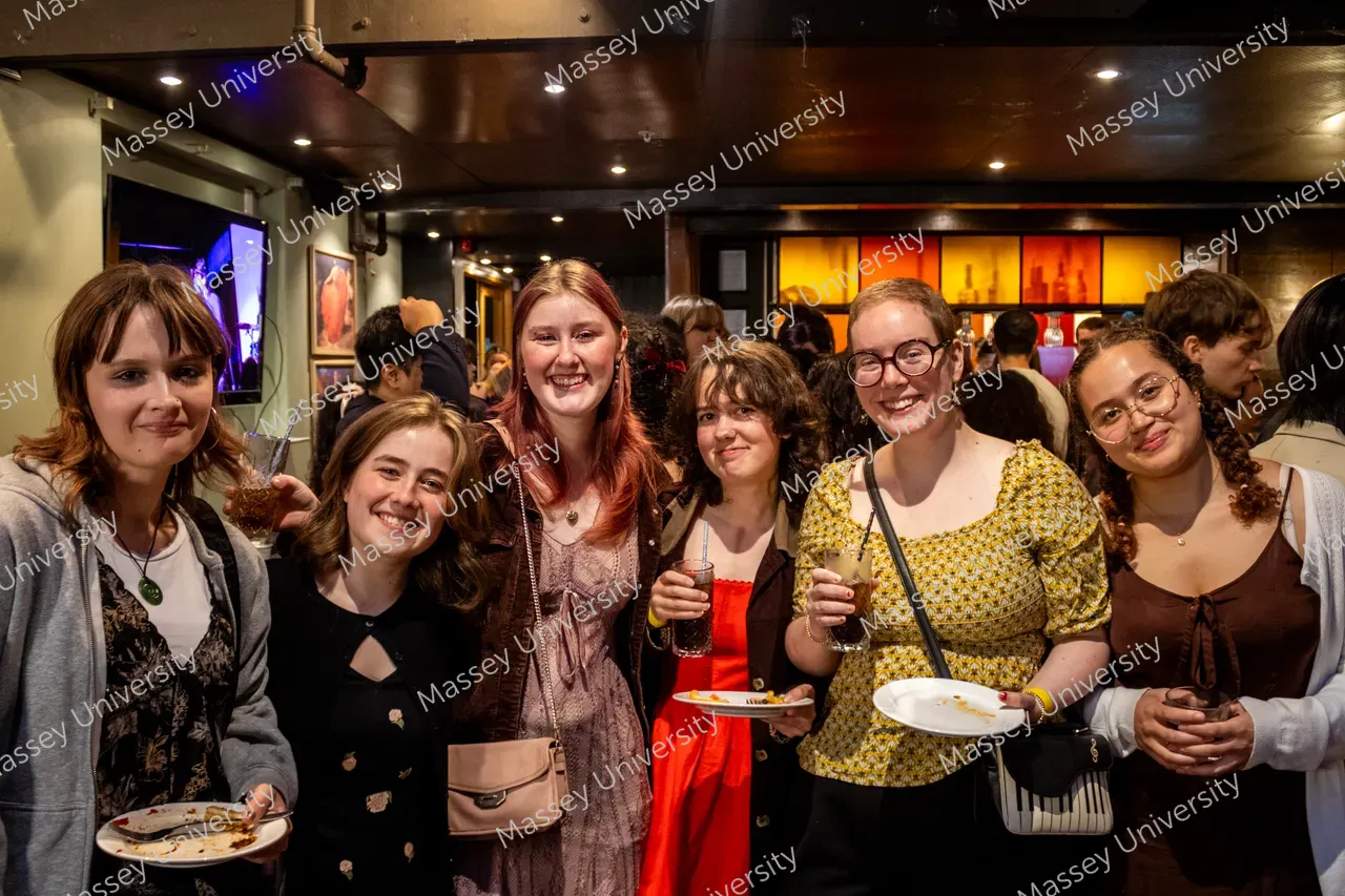 A group of students smile together at an indoor event, holding plates of food and drinks.