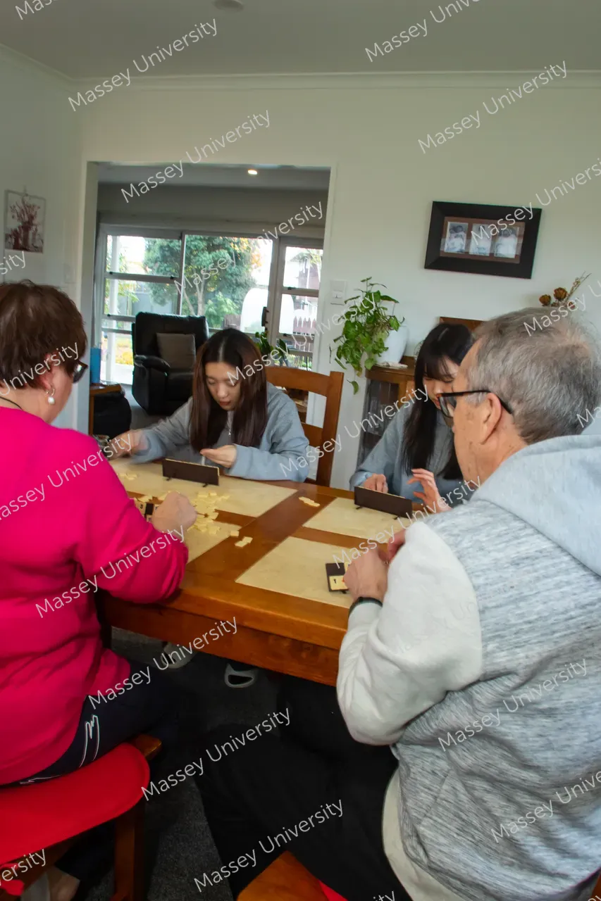 Four people play a tile-based game around a wooden table in a bright living room.